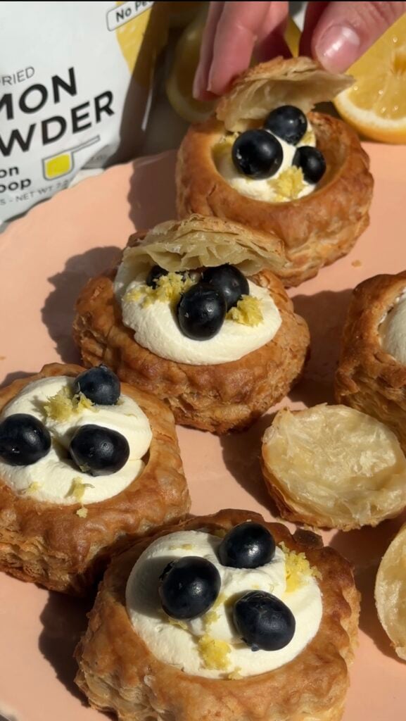 A hand placing a puff pastry shell filled with cream and topped with blueberries and lemon zest on a pink plate, surrounded by similar pastries. A lemon powder packet is partially visible in the background.