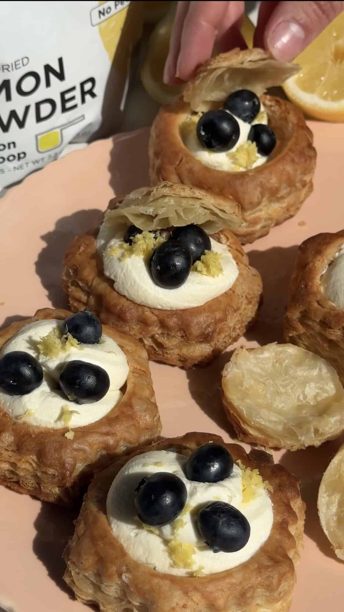 A hand placing a puff pastry shell filled with cream and topped with blueberries and lemon zest on a pink plate, surrounded by similar pastries. A lemon powder packet is partially visible in the background.