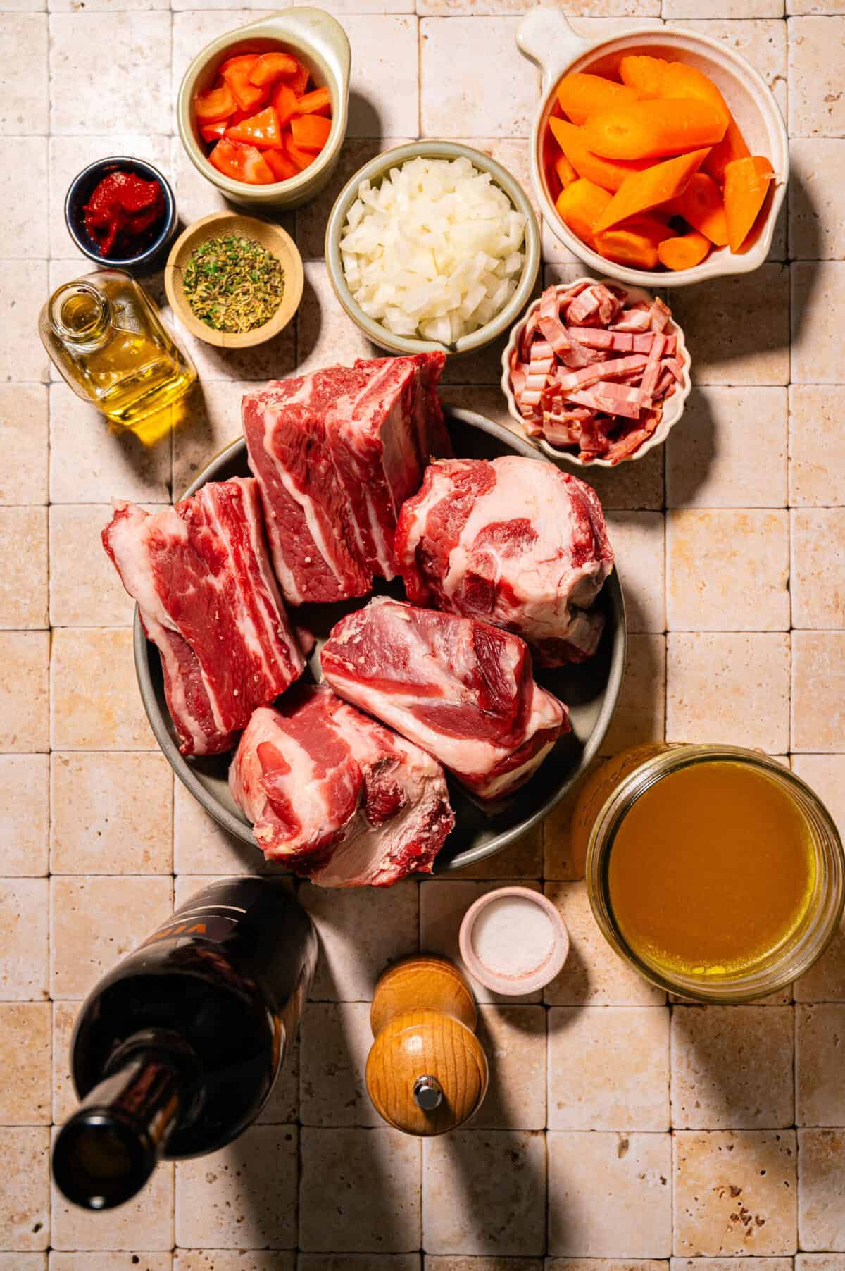 Raw beef short ribs, carrots, onions, bacon, chopped tomatoes, tomato paste, Italian herbs, olive oil, beef broth, salt, pepper, and a bottle of red wine arranged on a tiled countertop.