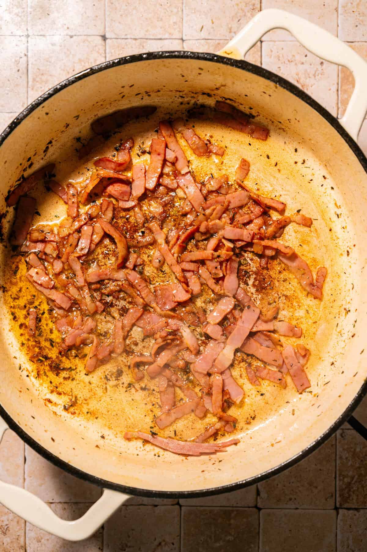 Sliced bacon is being cooked in a large cream-colored pot. The bacon pieces are browning and sizzling, and the pot has browned bits and oil on the bottom. The pot sits on a tiled surface.