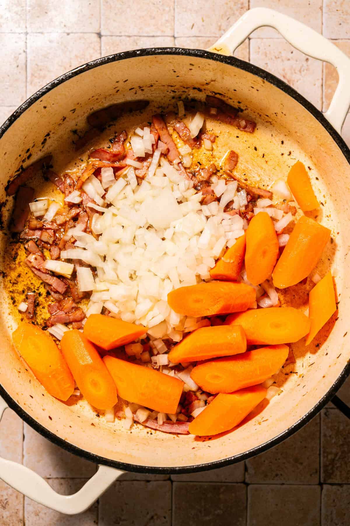 Chopped onions and large carrot pieces cooking with browned bits in a cream-colored pot on a tiled surface, viewed from above.
