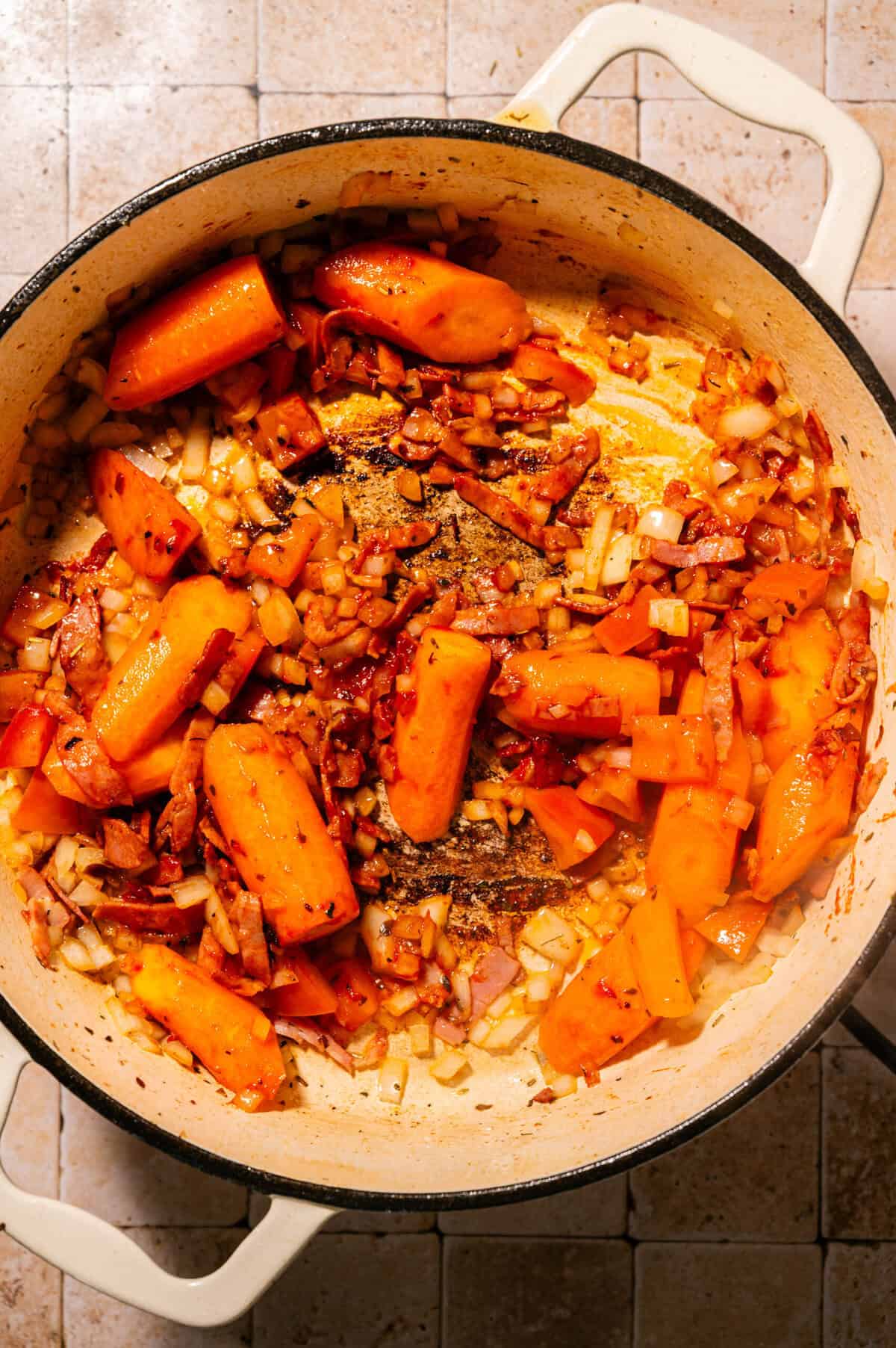 A white pot on a tiled surface filled with chopped onions, carrots, and tomatoes being sautéed, showing golden and slightly browned edges.