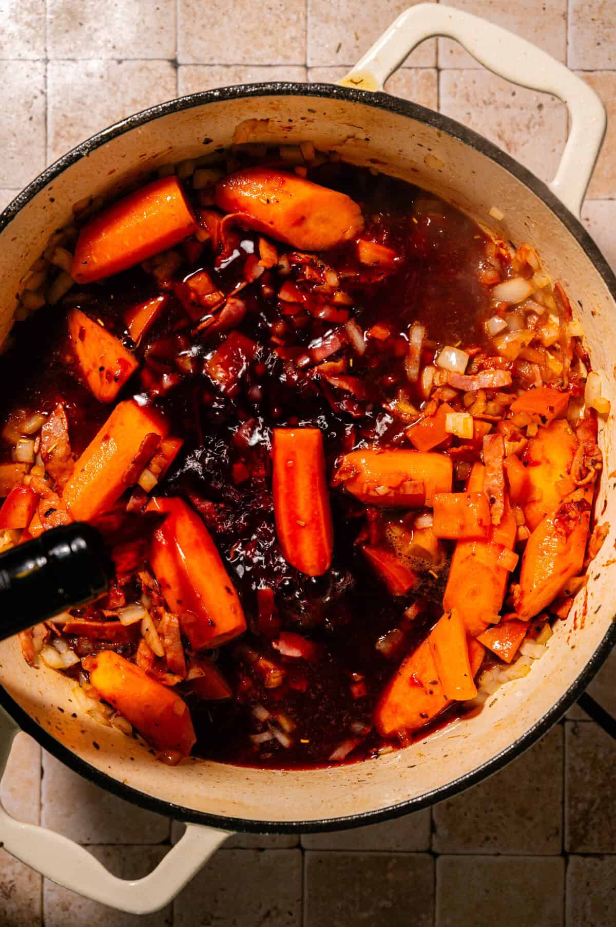 A pot filled with chopped carrots, onions, and meat simmering in a rich, dark sauce, viewed from above, on a tiled surface.