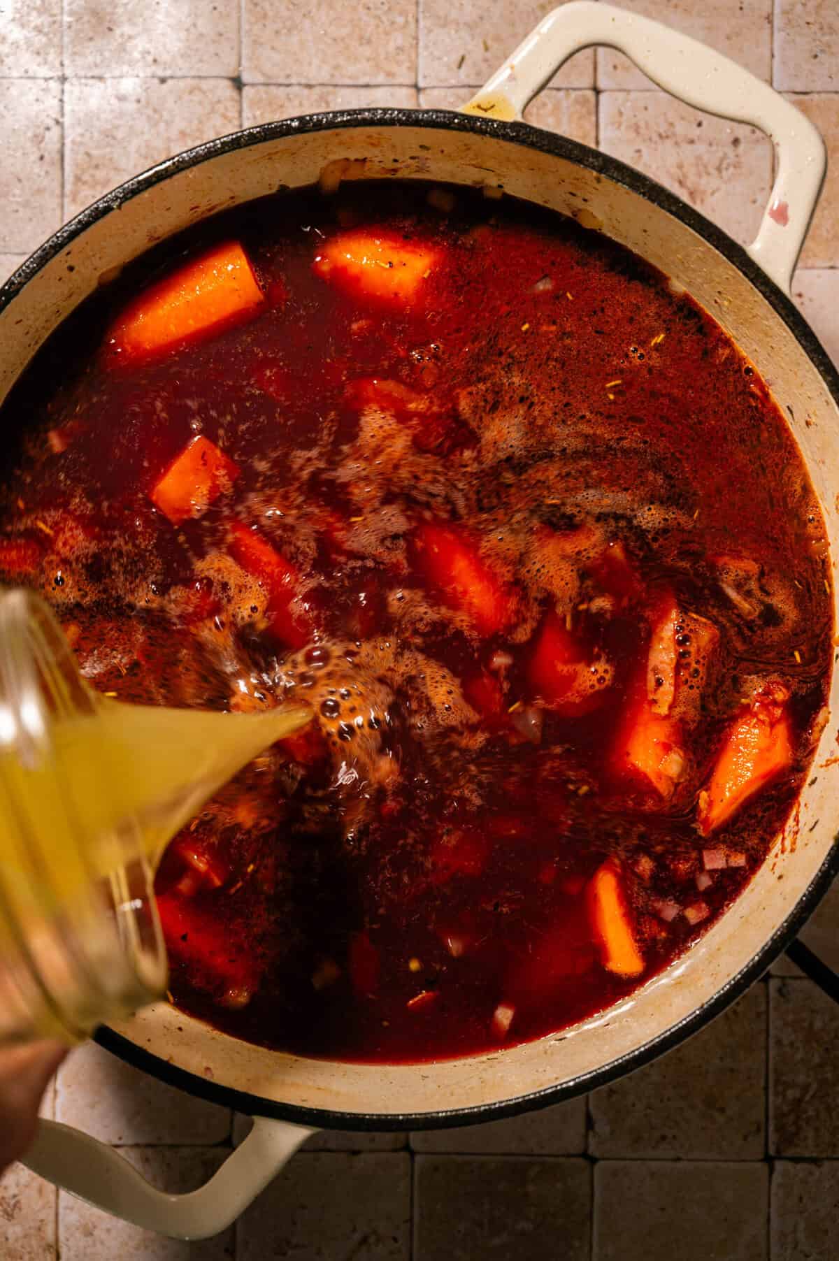 A pot of red soup with carrot chunks simmers as a yellow liquid is being poured in from a glass container. The pot sits on a tiled surface.
