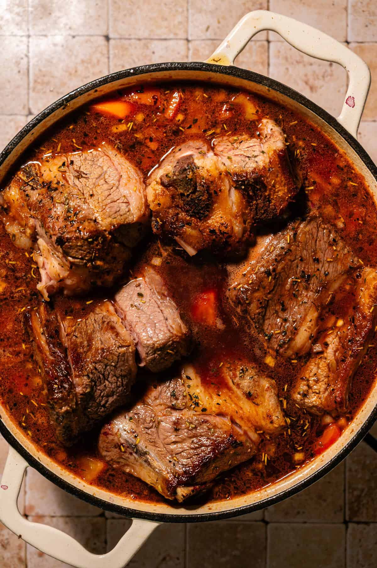 Large chunks of braised beef simmer in a rich, reddish-brown broth with herbs and vegetables, all in a white Dutch oven. The dish sits on a tiled countertop.