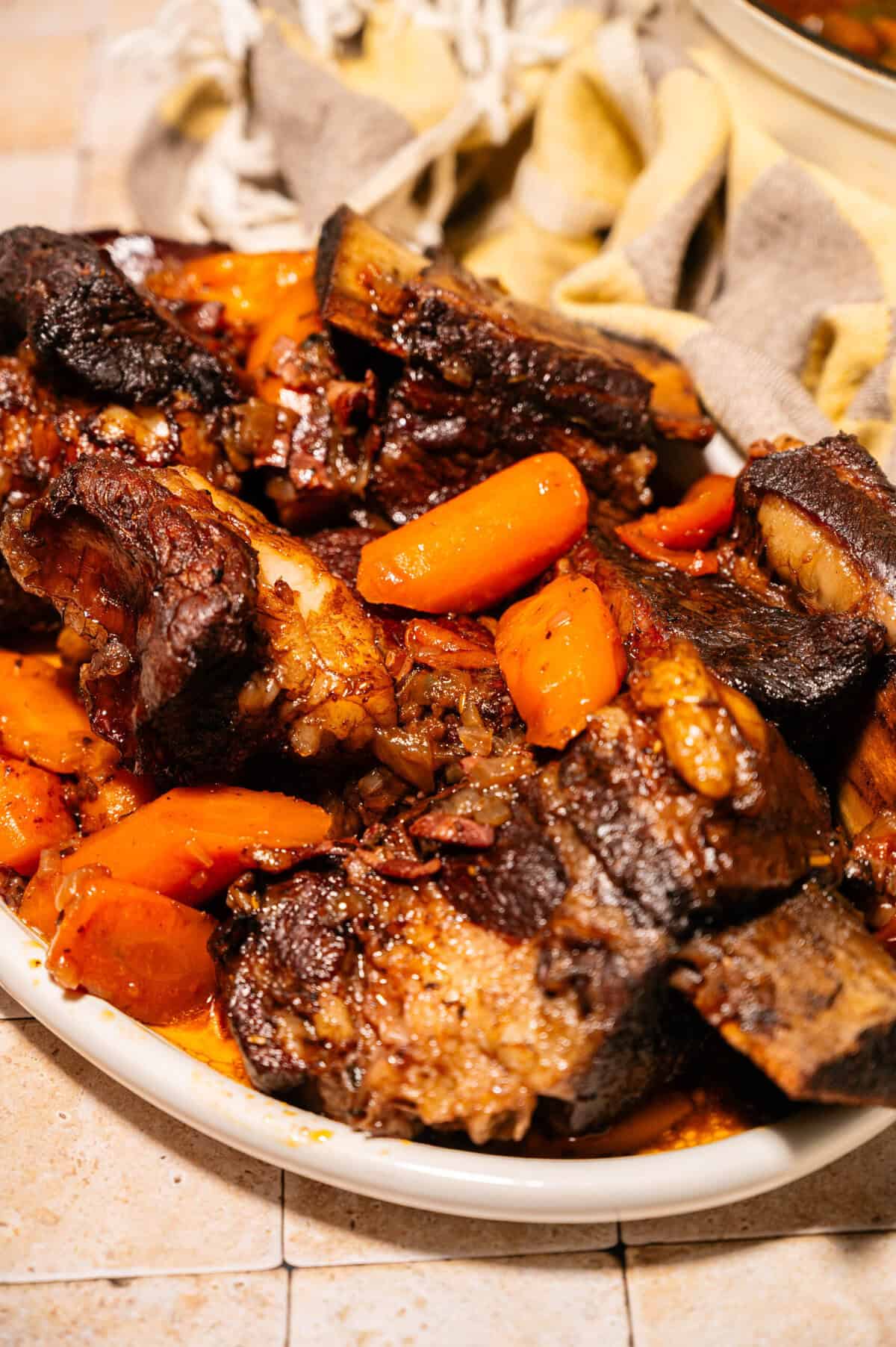 A close-up of braised short ribs with chunks of carrots on a white plate, garnished with sauce. A beige cloth is visible in the background.