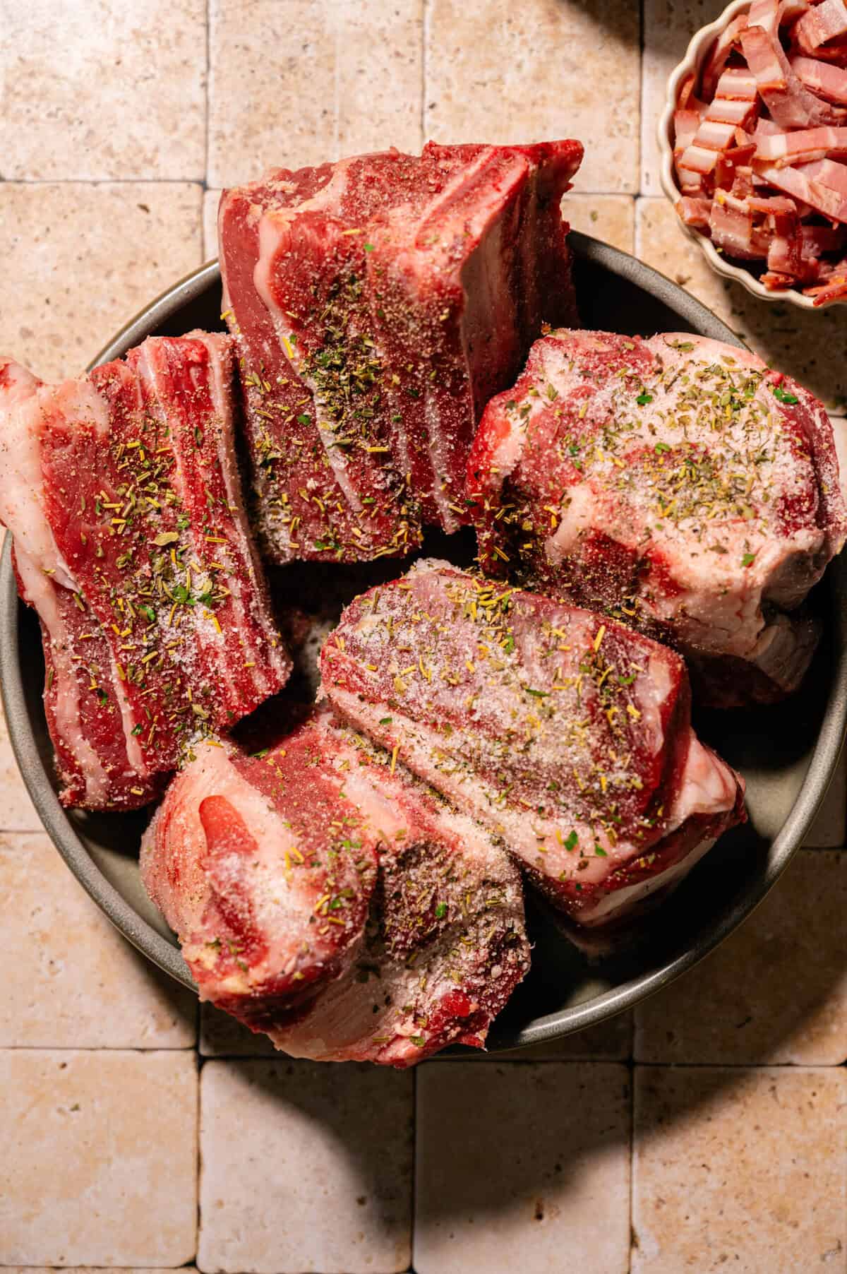 A bowl filled with raw, seasoned beef short ribs sits on a tiled surface; a small bowl of diced pancetta is visible in the corner.