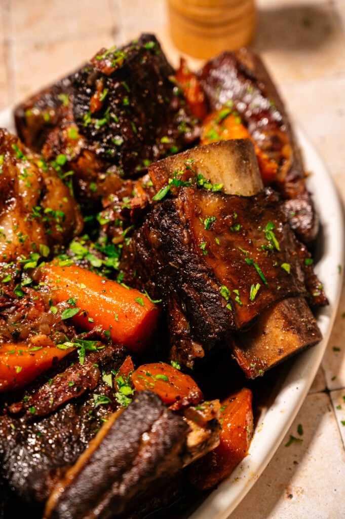 A close-up of braised beef short ribs with carrots, garnished with chopped parsley, served on a white plate.