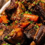 A close-up of braised short ribs with carrots, garnished with chopped parsley, served in a white dish. The meat appears tender and richly browned, surrounded by a savory sauce.