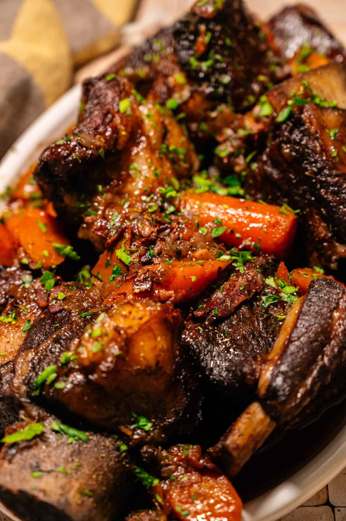 A close-up of braised short ribs with carrots, garnished with chopped parsley, served in a white dish. The meat appears tender and richly browned, surrounded by a savory sauce.