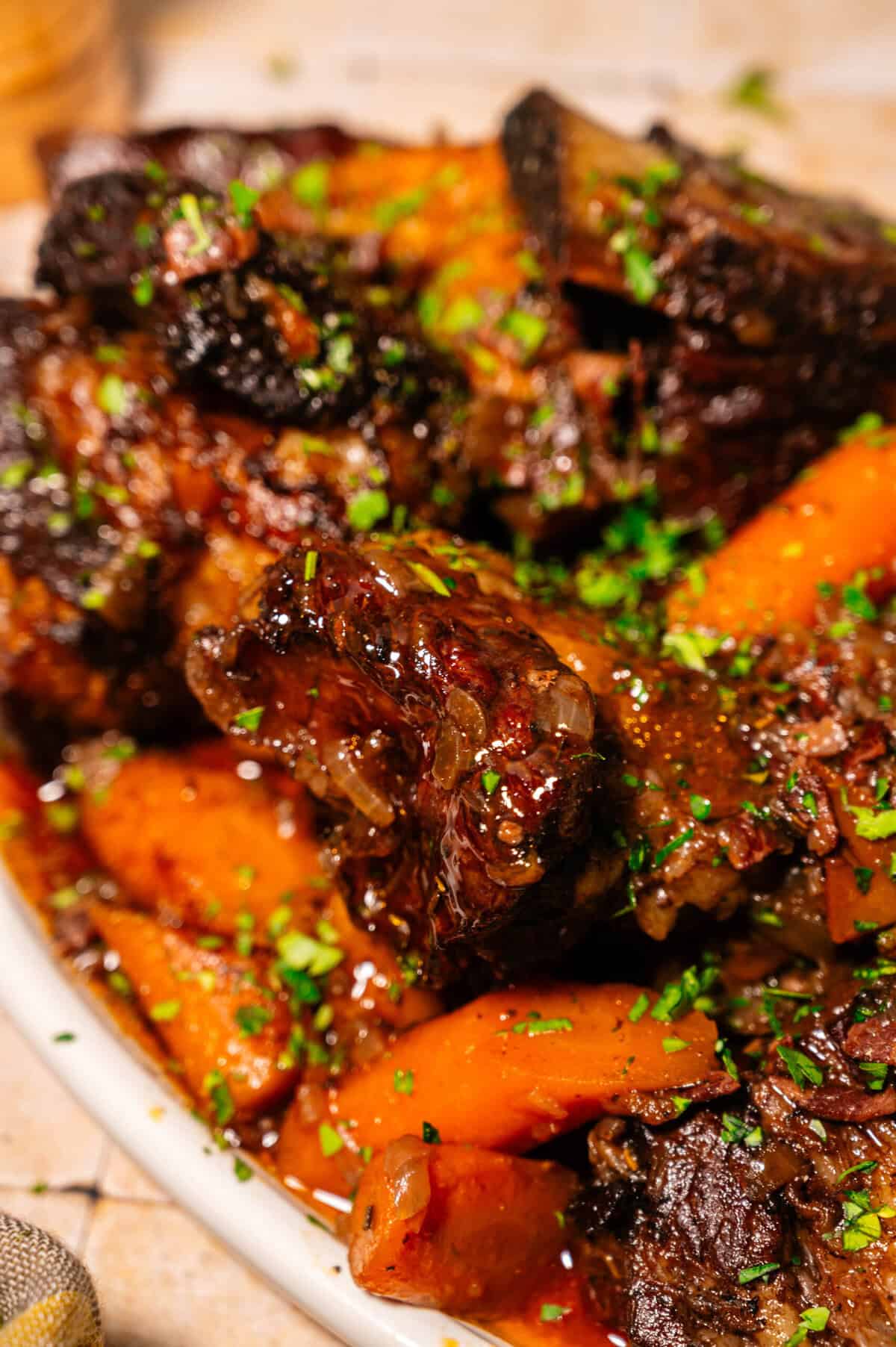 Close-up of braised short ribs with carrots, garnished with chopped parsley, served on a white plate. The meat looks tender and richly glazed.
