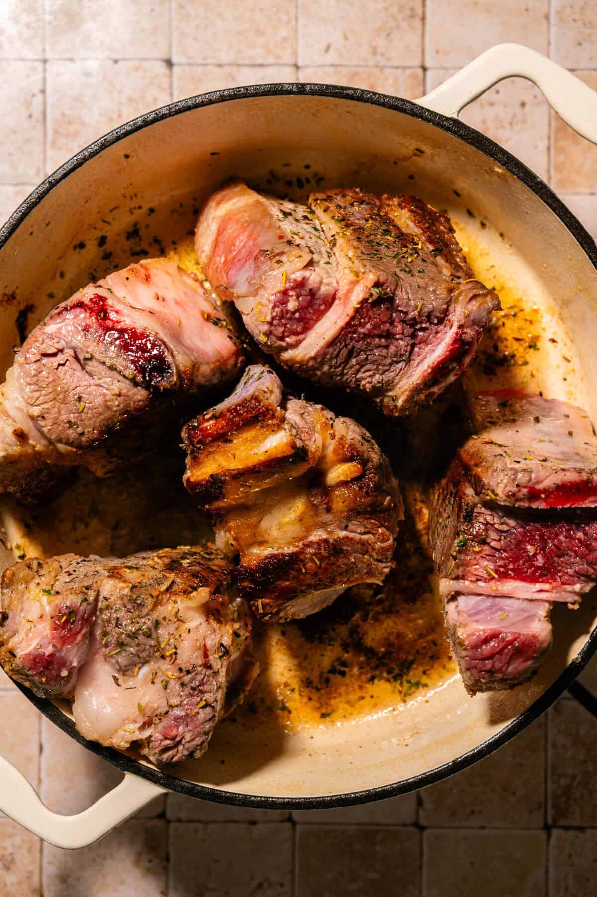 Large pieces of browned beef short ribs searing in a round, white Dutch oven, with visible seasoning and rendered fat, on a tiled countertop.