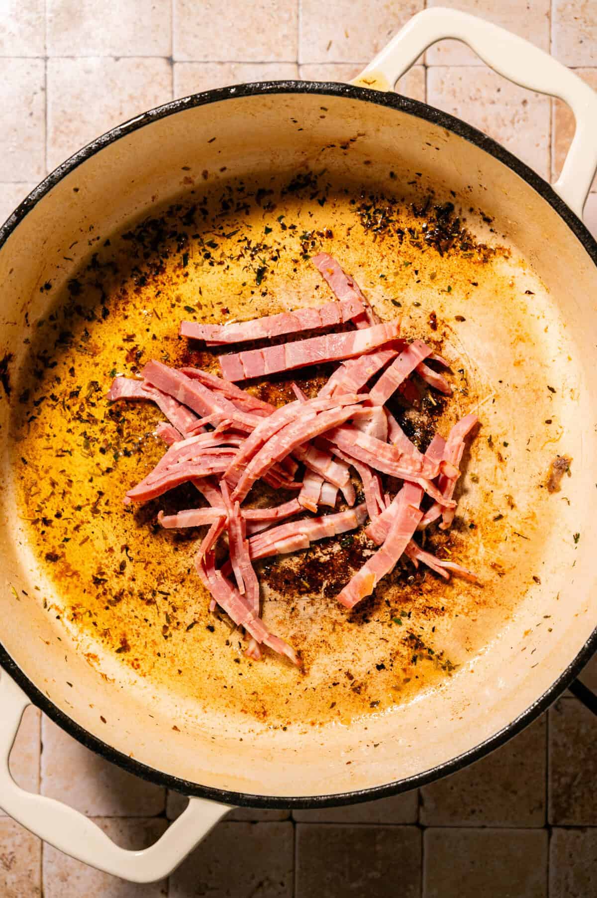 Sliced bacon is cooking in a large, white enameled pot with browned bits and seasoning on the bottom, sitting on a tiled countertop.