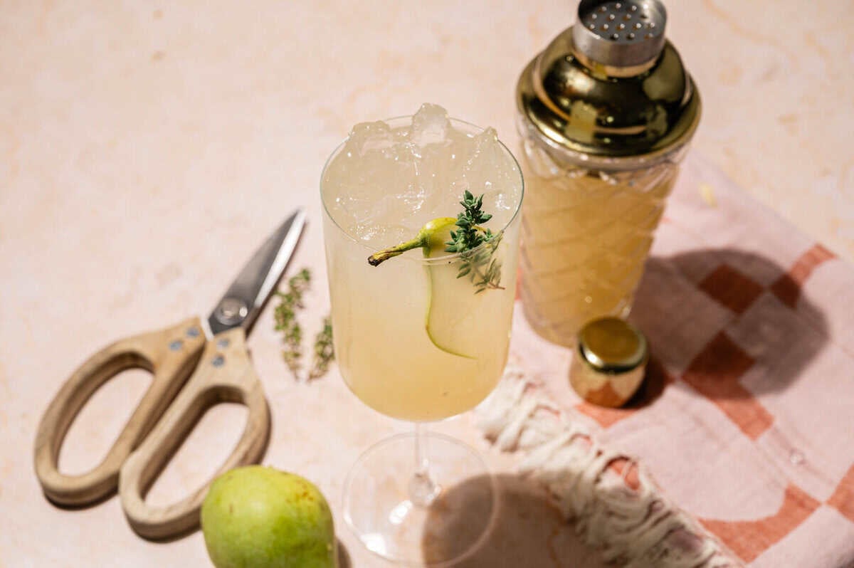 Pear Collins cocktail on a pink table with kitchen shears, fresh thyme, and a whole pear