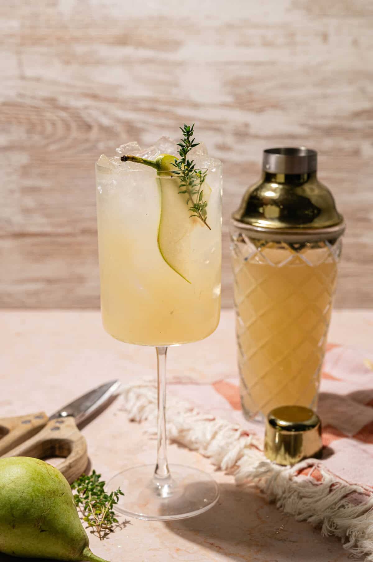 Pear cocktail garnished with a thin pear slice and fresh thyme next to a gold cocktail shaker