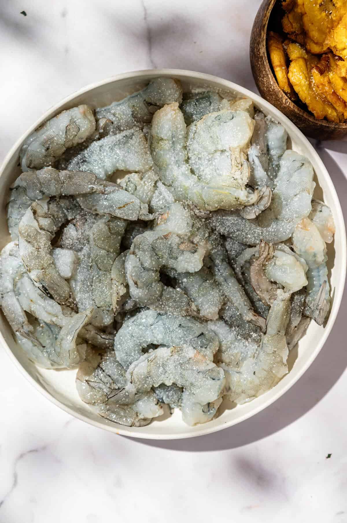 Raw peeled shrimp seasoned with salt in a white bowl for encocado de camarón