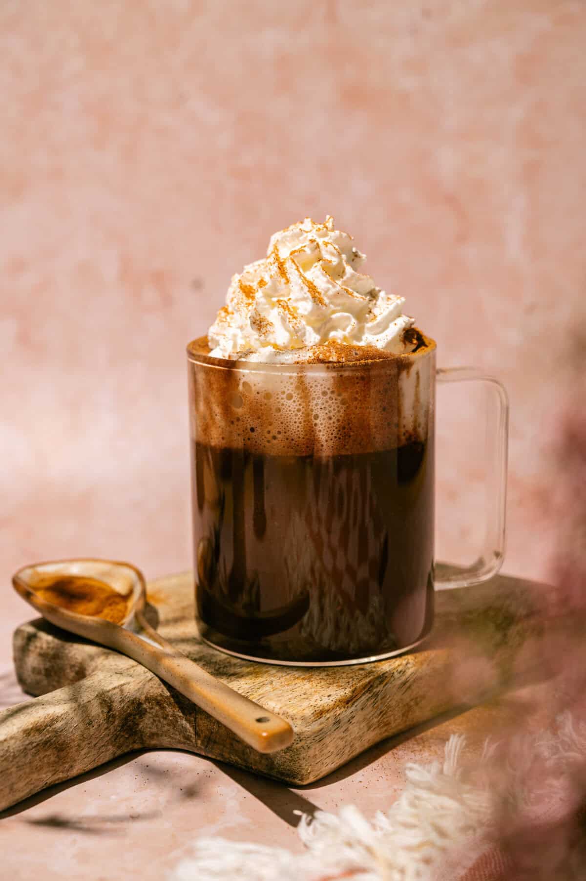 Glass mug of creamy hot chocolate with a foamy top and whipped cream dusted with cinnamon on a wooden board.