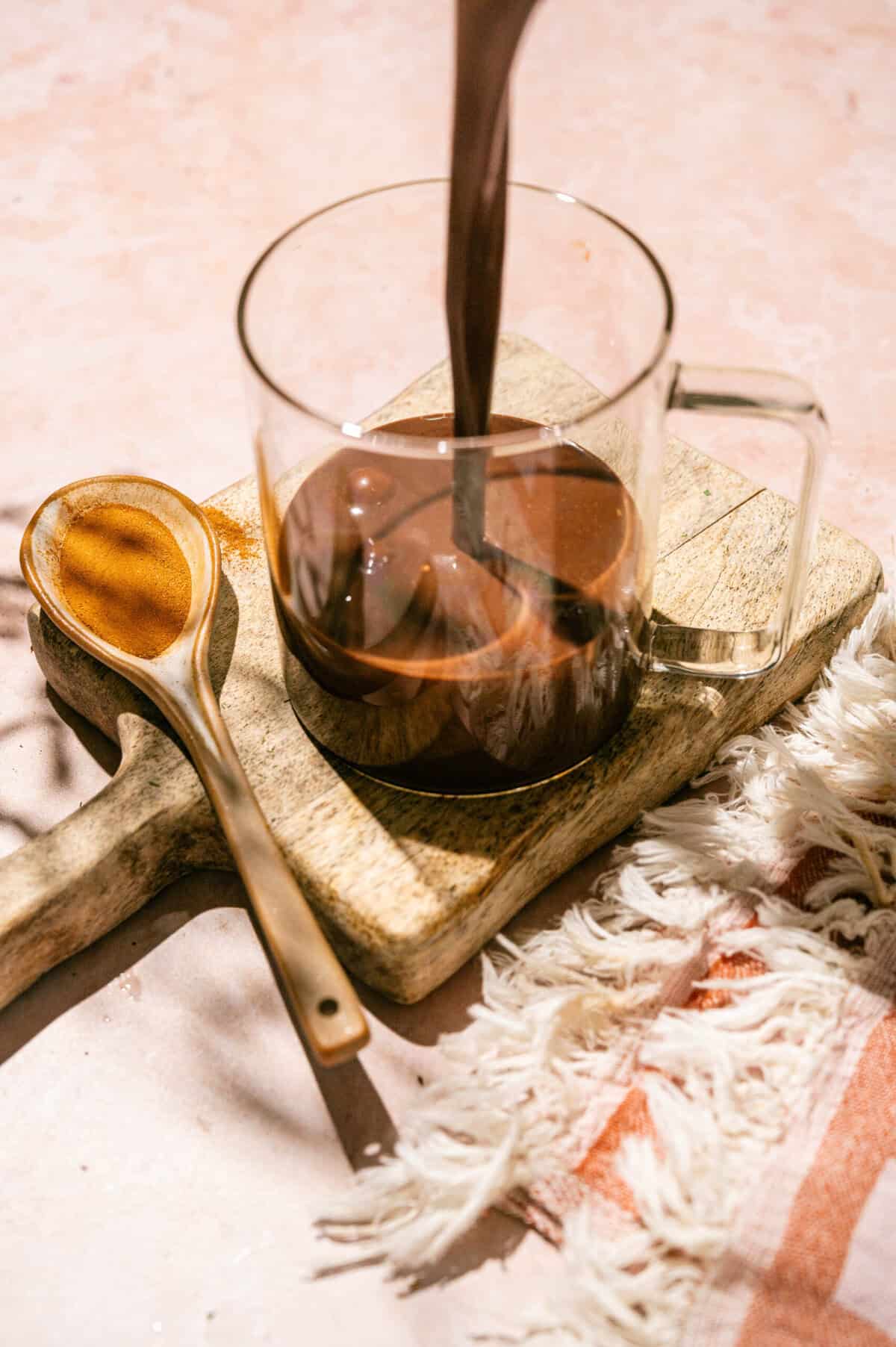 Pouring rich homemade hot chocolate into a clear glass mug on a wooden board next to a ceramic spoon of cinnamon.