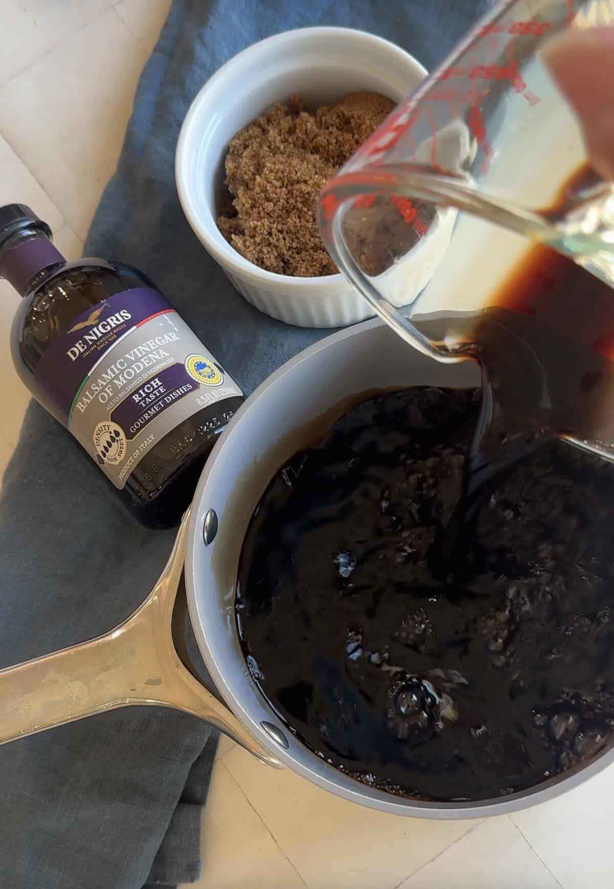 Balsamic vinegar poured from a glass measuring cup into a saucepan, with a ramekin of brown sugar nearby