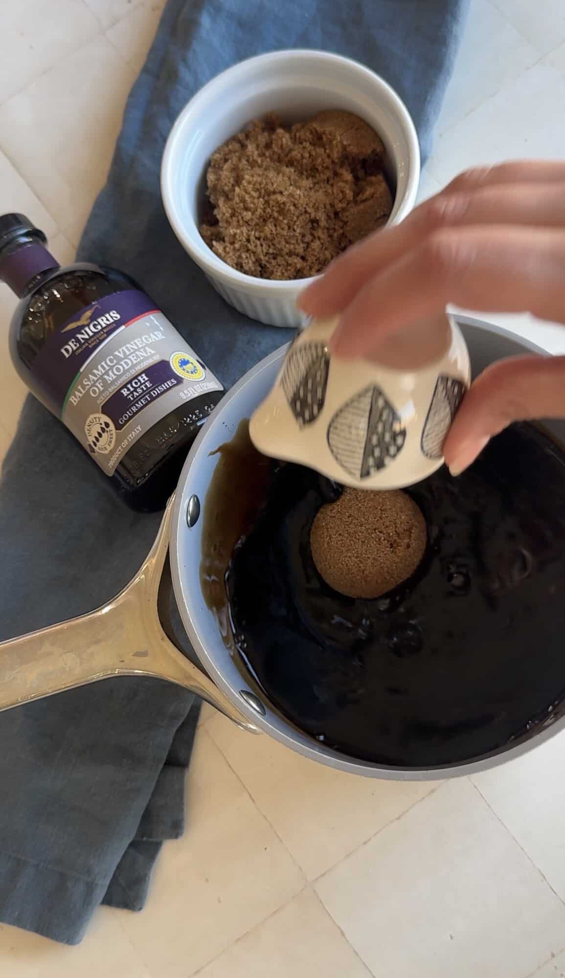 Hand adding brown sugar to a saucepan of balsamic vinegar to make balsamic glaze