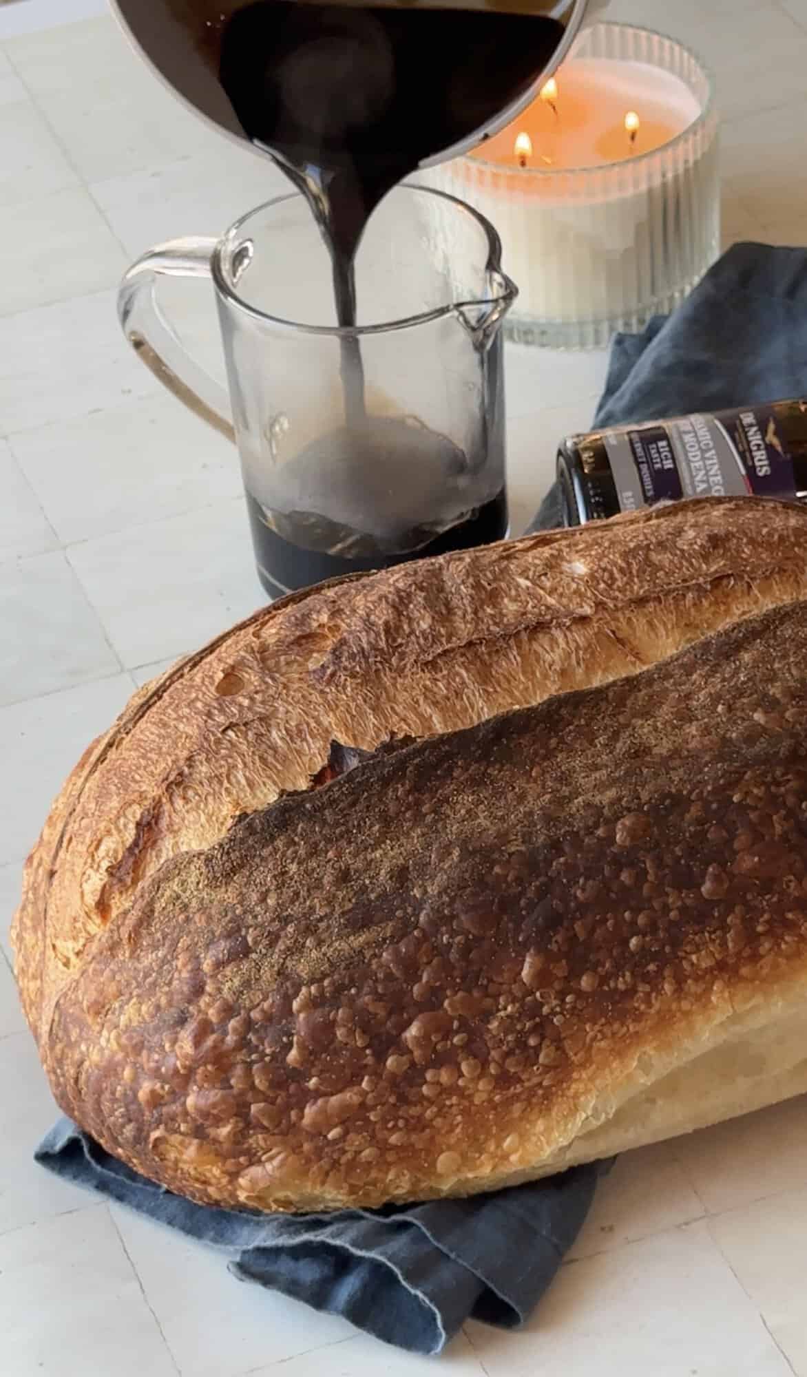 Hot balsamic glaze poured from a saucepan into a glass pitcher next to a loaf of sourdough bread