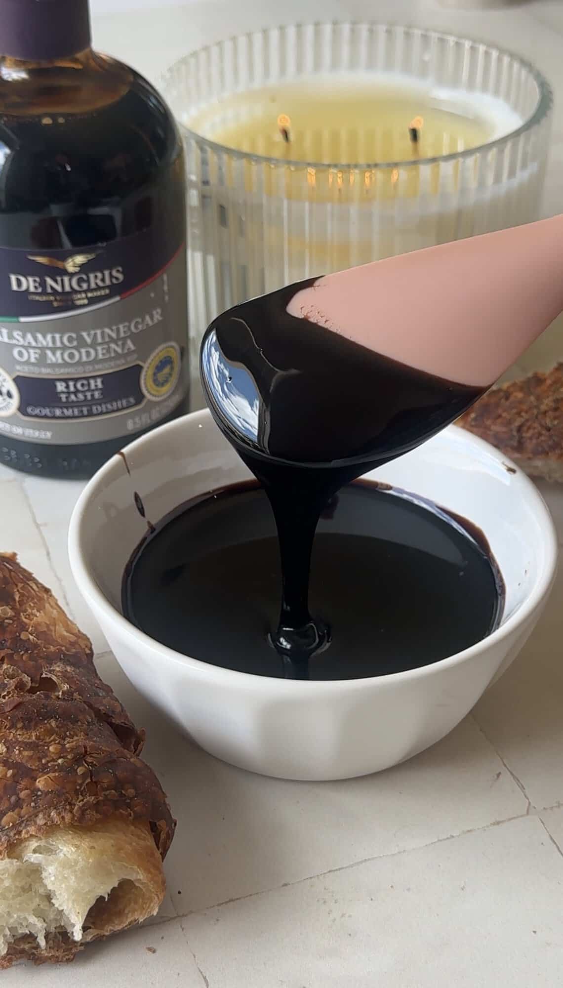Thick balsamic glaze dripping from a pink spoon into a small white bowl, with a De Nigris balsamic vinegar bottle behind it