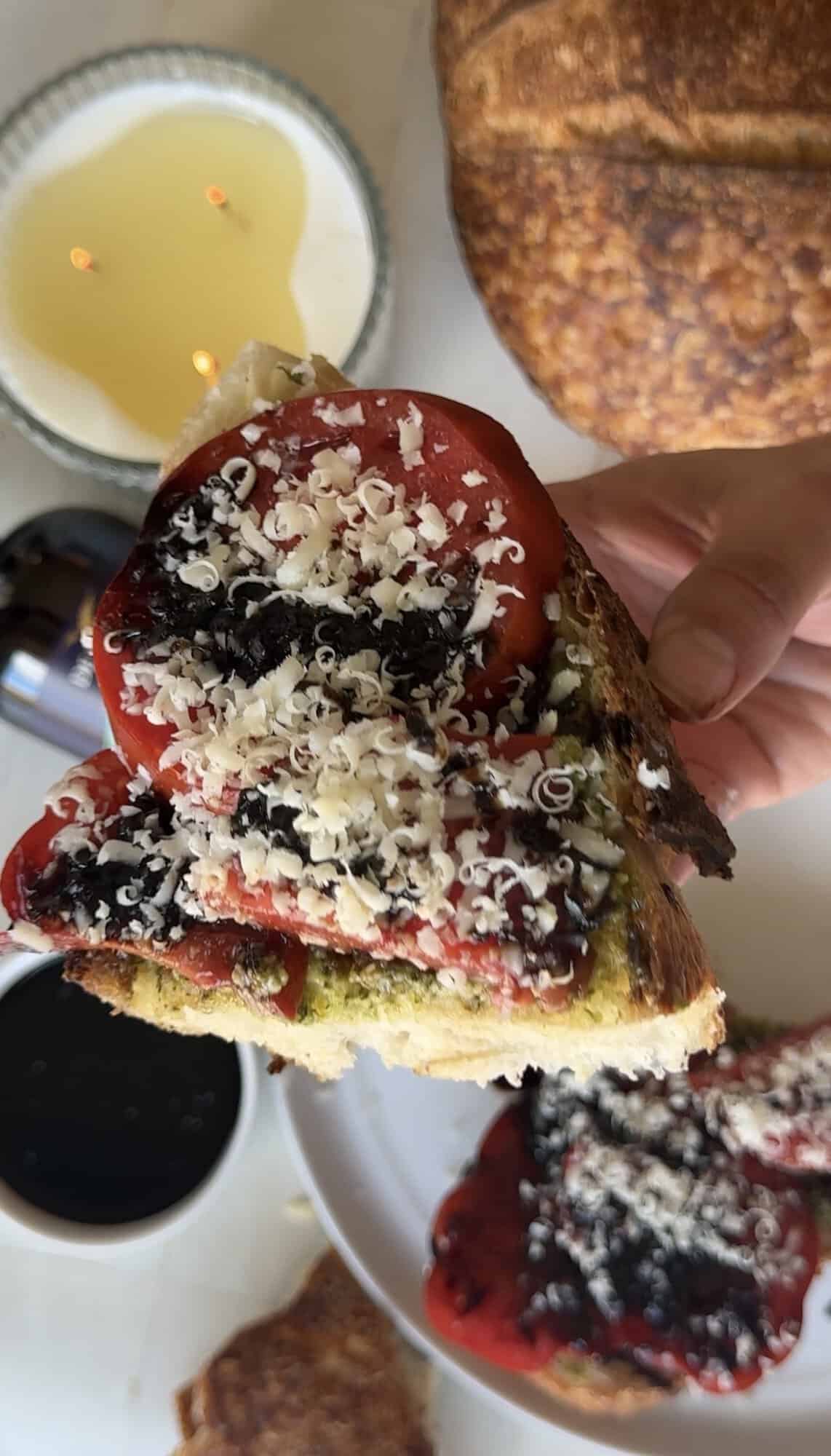 Hand holding sourdough toast topped with pesto, sliced tomatoes, balsamic glaze, and freshly grated parmesan