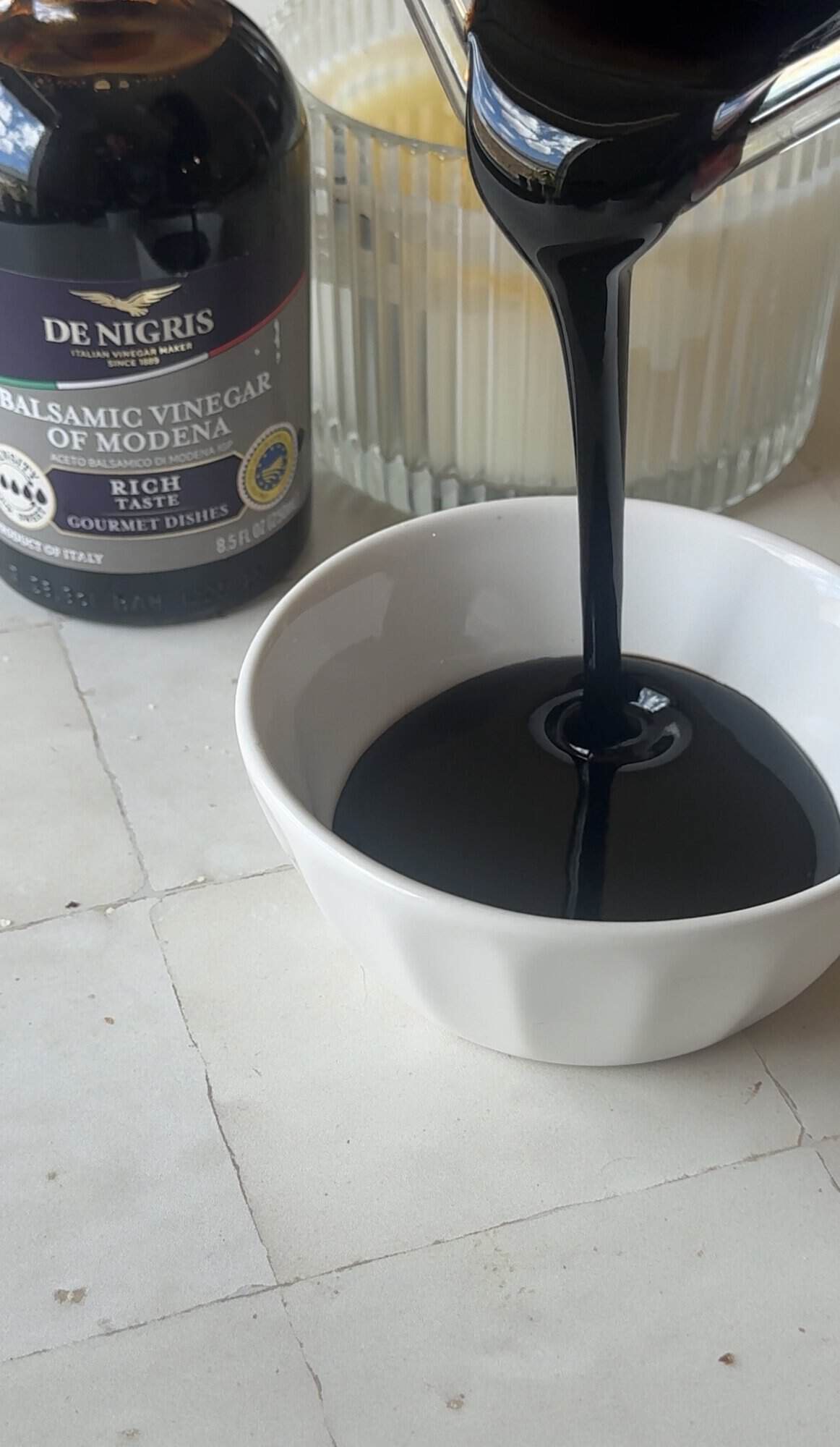 Thickened balsamic glaze poured from a saucepan into a small white bowl on a tile countertop
