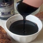 Thick balsamic glaze dripping from a pink spoon into a small white bowl, with a De Nigris balsamic vinegar bottle behind it