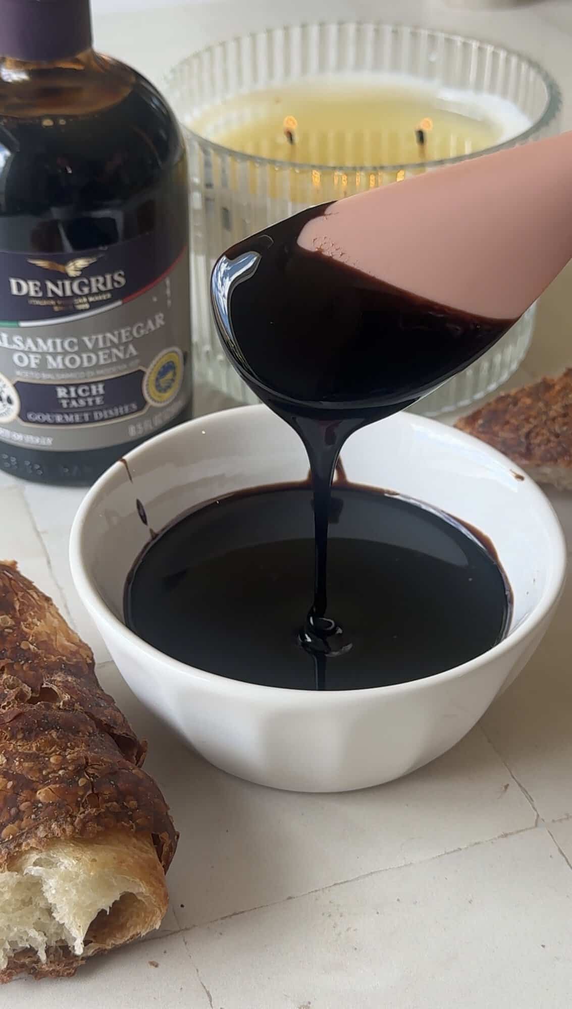 Thick balsamic glaze dripping from a pink spoon into a small white bowl, with a De Nigris balsamic vinegar bottle behind it