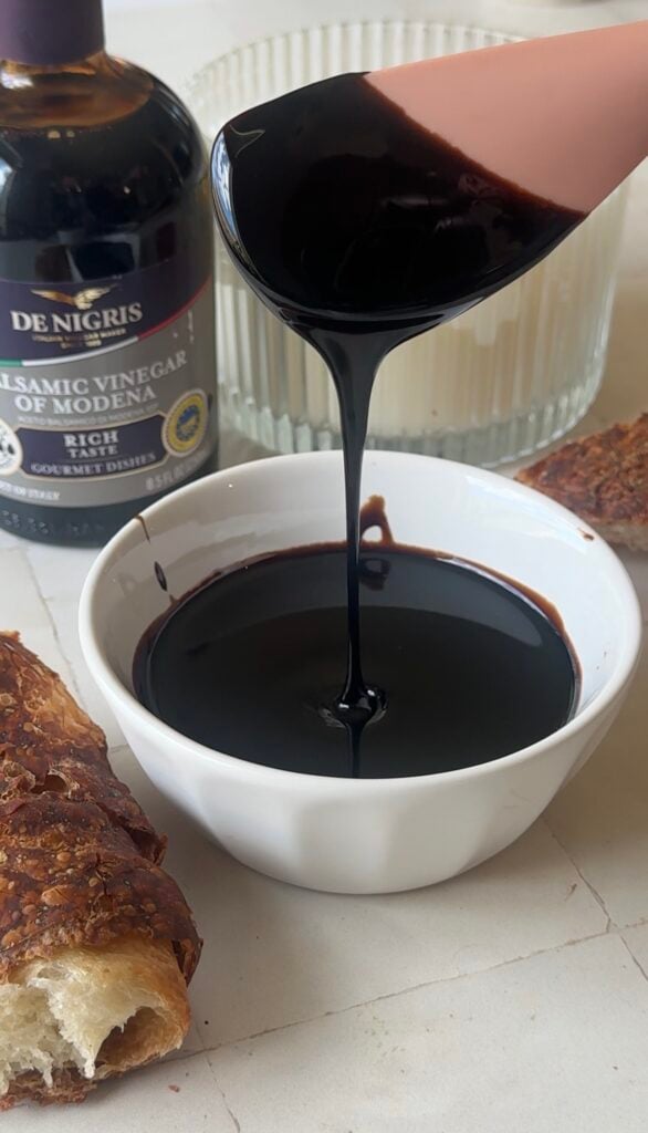 Thick homemade balsamic glaze dripping from a spoon into a white bowl, showing its glossy, syrupy texture