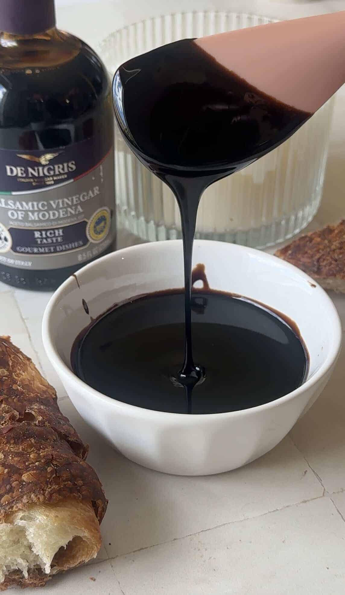 Thickened balsamic glaze poured from a saucepan into a small white bowl on a tile countertop
