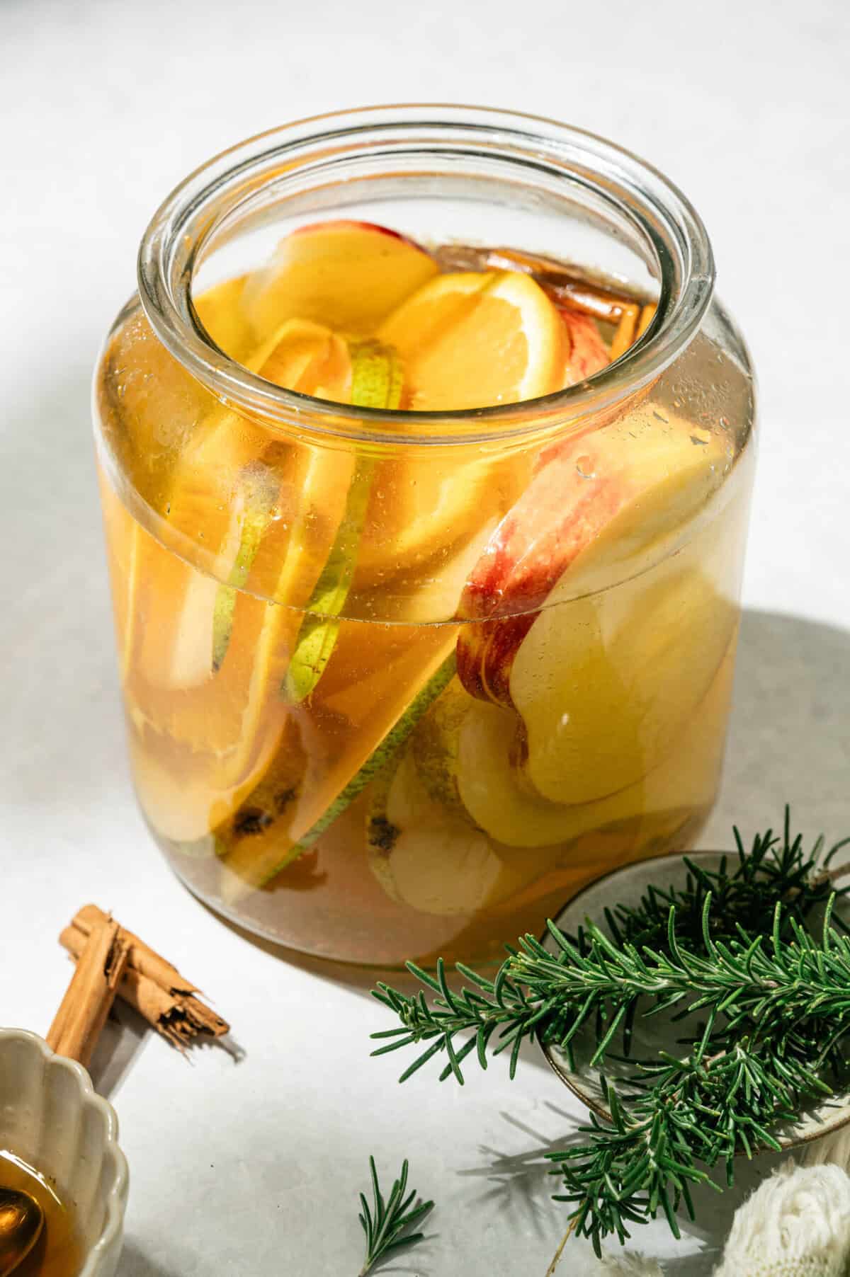 Glass jar of apple cider sangria with pears, apples, oranges, and cinnamon sticks next to fresh rosemary