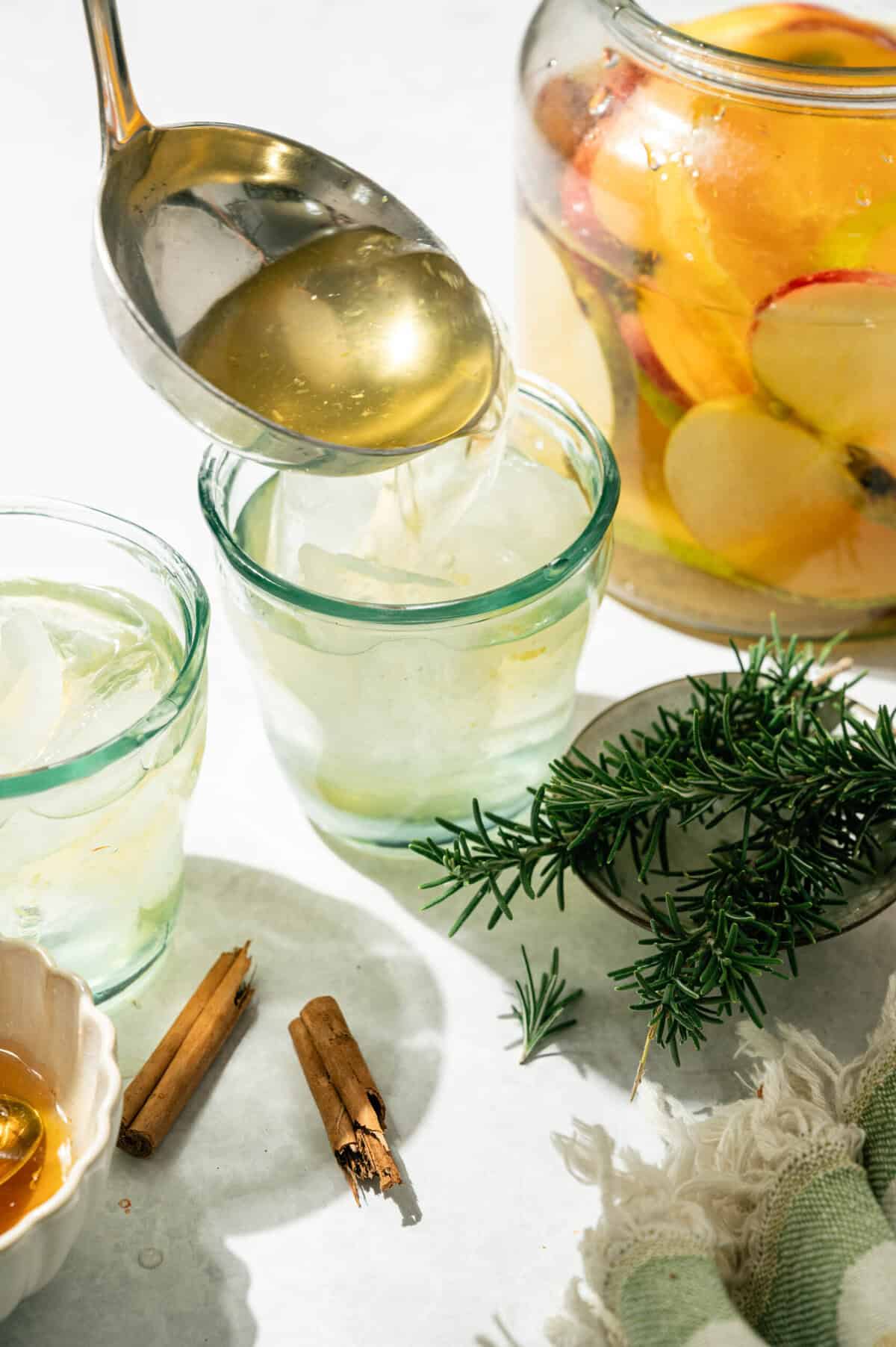 Ladling apple cider sangria into a glass over ice with a jar of fruit, rosemary, and cinnamon sticks nearby