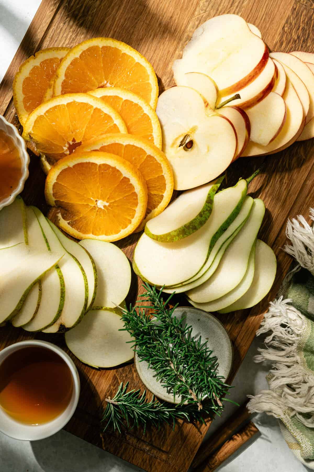 Sliced oranges, Honeycrisp apples, and pears on a cutting board with rosemary and honey for apple cider sangria