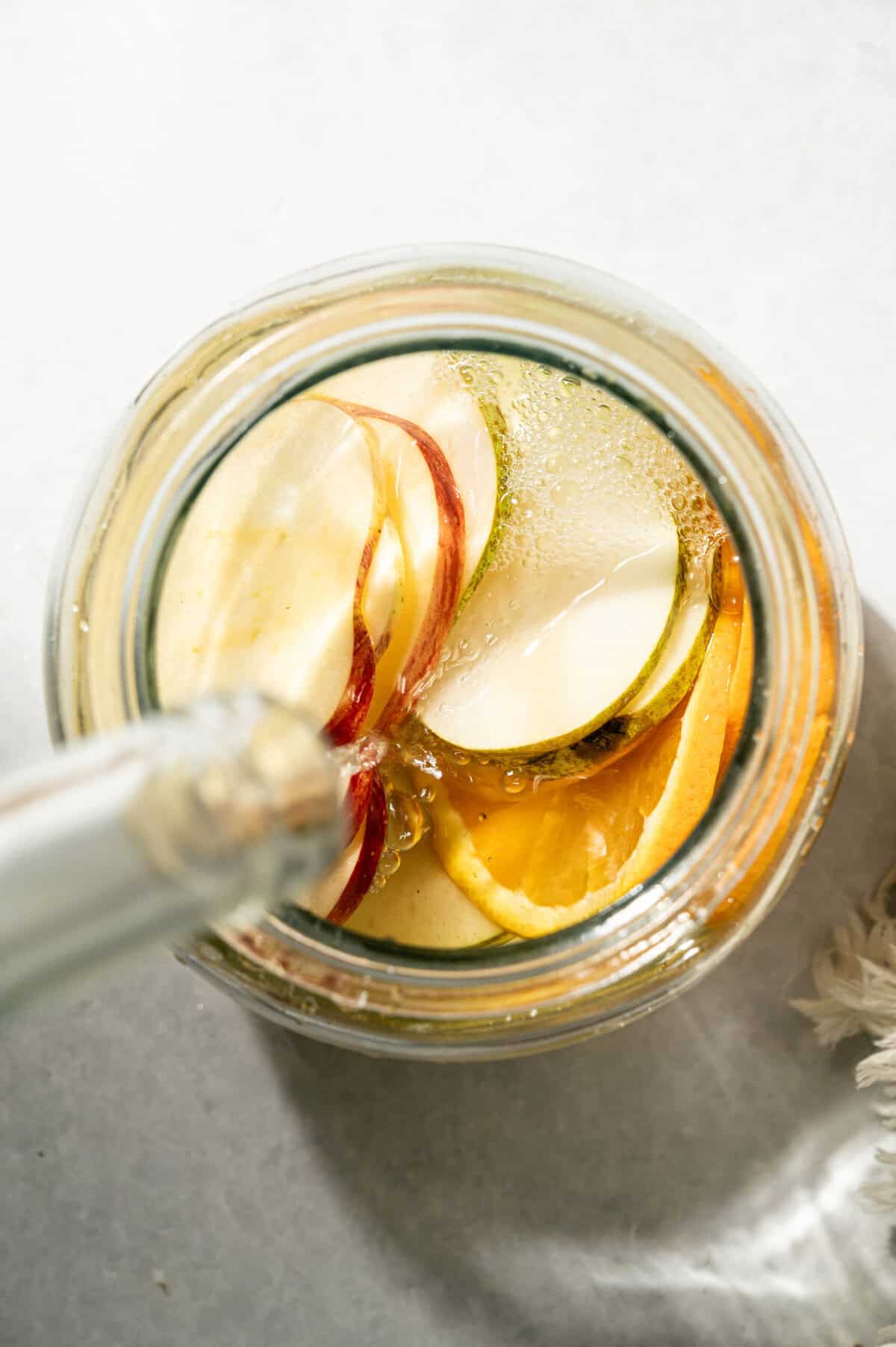 Pouring white wine into a glass jar of apple, pear, and orange slices to make apple cider sangria